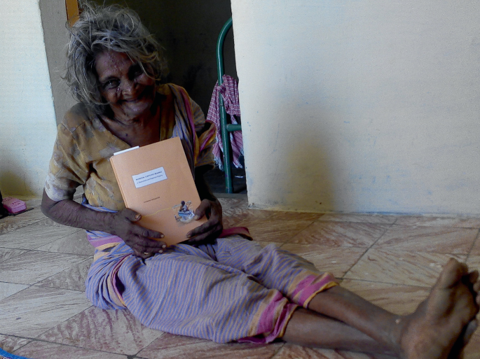 A photograph of an old woman with a thesis in her hands. The woman sits on the floor.