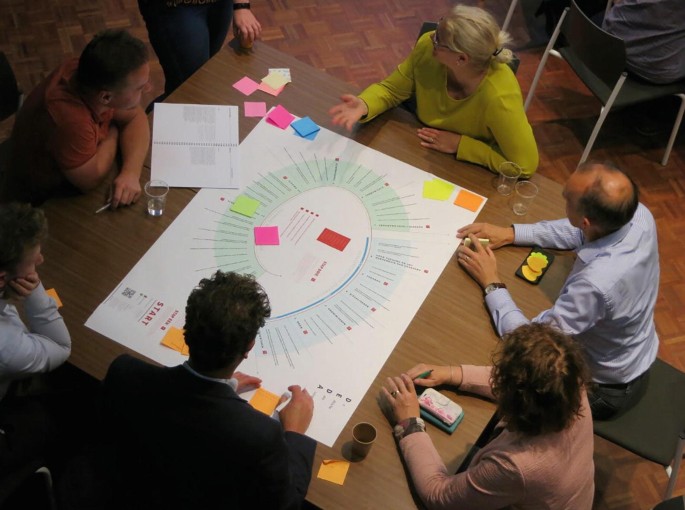 A photograph. A group of 6 people is seated around the table. A project chart is placed on the table.