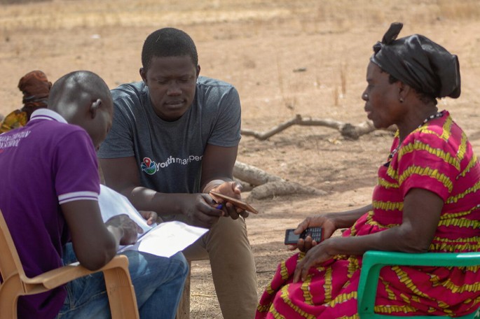 A photograph of 3 people sitting on chairs in a round manner. Two men and an elderly woman where the young man checks some details on the notes.