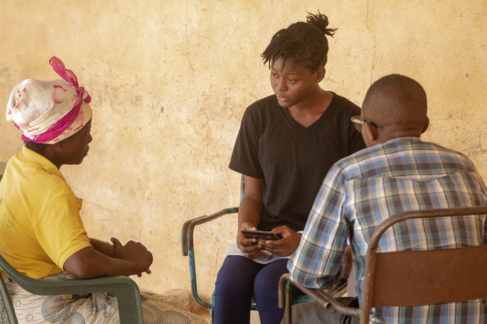 A photograph depicts 3 people sitting on chairs in a round manner. Two members of the Youth Mappers team collect data from an elderly woman.