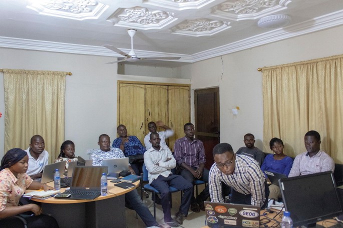 A photograph of a group of people in a presentation room. A few of them look at the laptops placed in front of them, and others sit at the back.