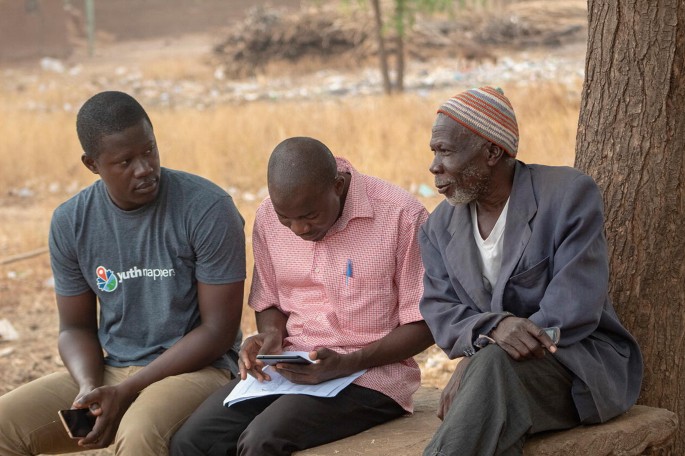 A photo depicts 3 men sitting under a tree. The author and a man collect data from an elderly man.