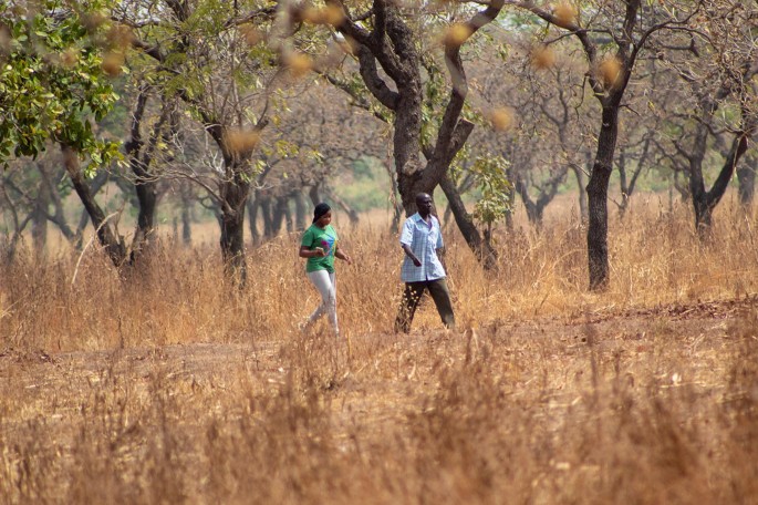 A photo depicts a woman and a man walking in a forest, that has dried grass and trees. The woman walks behind the man carrying a mobile in her hand.