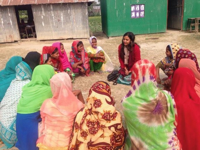 A photograph depicts a group of women sitting on the ground and interacting with a Youth Mapper. There are rows of shed houses behind them.