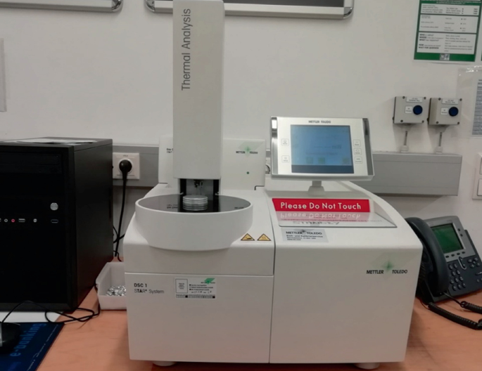 A photo of a differential scanning calorimetry device. The device has a digital output screen on the top. Below which a sign board reading, please do not touch is placed. A vertical part is placed to the left of the screen. All these parts are placed on the base divided into 2 compartments