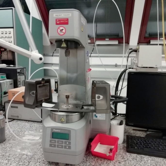 A photo of rotational rheometer. The device has a cabinet above, with a vertical protrusion, which is the sampling space. The controls, and the readings are present below the sampling area. The device is connected to stabilizers and screens. Narrow pipes run to and from the device.