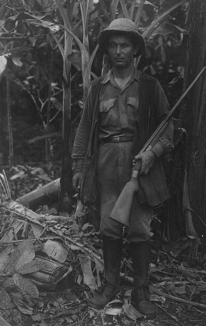 A photograph of Jose Correia standing in the woods, wearing hunting clothes, during his bird-collecting mission. He has a shotgun in his hands.