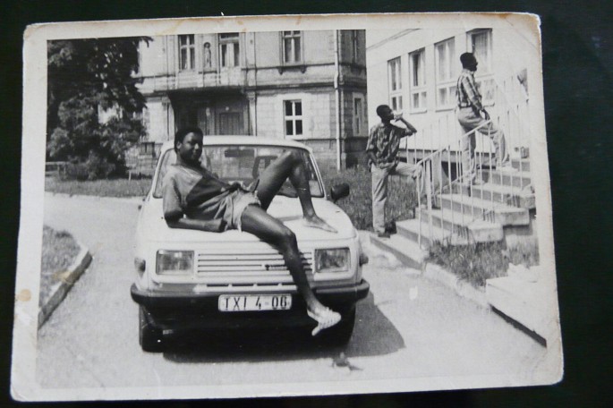 A photograph of a man half lying on the bonnet of the car and two persons stand on staircase of a house.