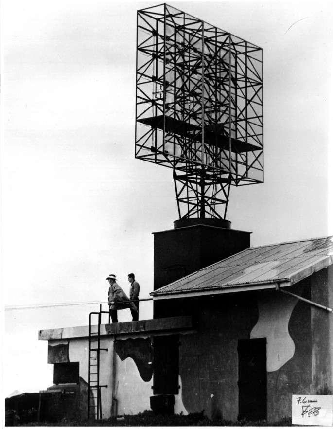 An old monochrome photograph of a square-shaped metal frame with a gridded framework is placed on an elevated platform. Two men stand at the foot of the structure.