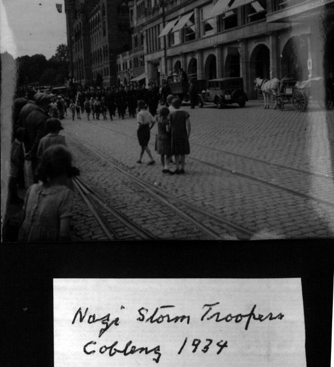 A photograph of Nazi troopers as they parade down the road with onlookers on the sides.