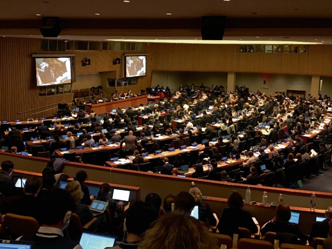 A picture of a large conference room includes a large group of people seated, each with a laptop open. It depicts the person speaking on the two big screens.