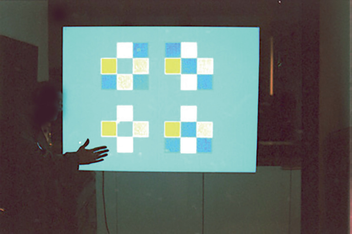 A photo of an interactive video construct at Kettles Yard in Cambridge portrays a man demonstrating formal systems to make interactive generative art. On the screen are seen several shapes created with white, blue and yellow squares.