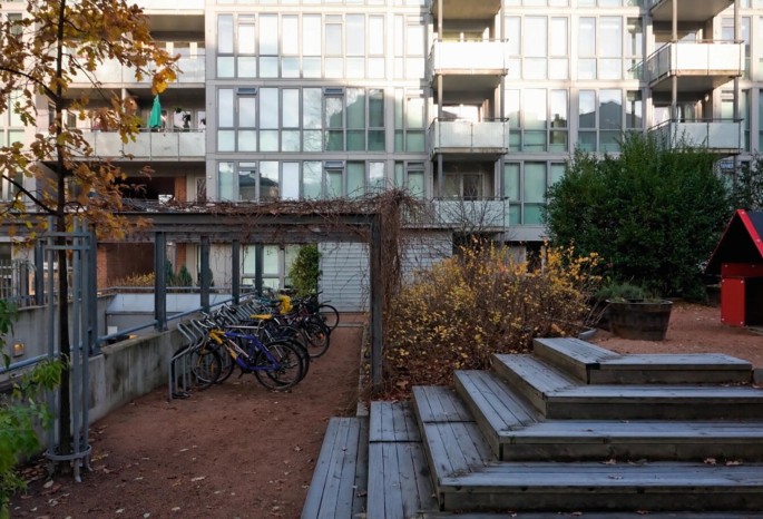 A photograph of the glass-windowed fa&#x00E7;ade of Klosterenga viewed from a garden with a short wooden stair on the right and bicycles parked on the left.
