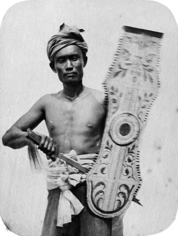 A photograph of an indigenous person carries armour and a shield.