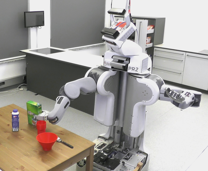An photograph of a P R 2 robot performing a task of setting a table. The robotic arm holds a box package placed on a table.