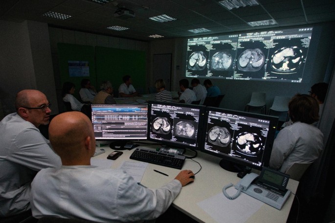 A photo of a team of tumor board members discusses at a workstation, a screen in of them displays x-ray images.