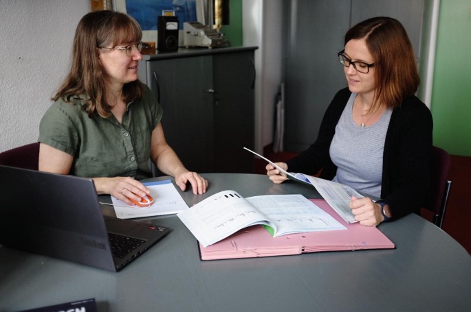 A photograph of two women sitting around a table in a room. One of them glances at the report that she holds. While the other woman places her right hand on the mouse of a laptop in front of her.