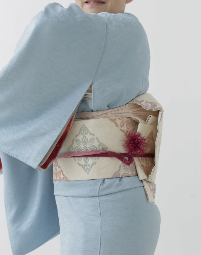 A photograph of a woman, who wears a light-colored denim dress with bright fabric around the waist.