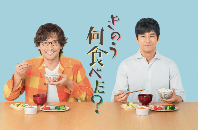 A photograph of 2 men sitting in front of a table, and holding a bowl, and a chopstick in their hands. There are some food items lying on the table. Some text is provided in a foreign language.