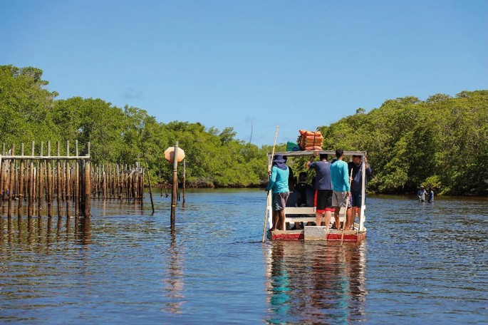 A photo of the Jangada boat in the water. Few people stand behind the boat, and few of them sit inside.