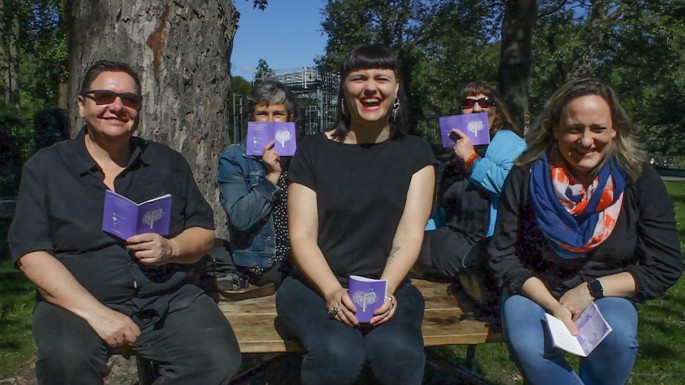 A photograph of five people sitting under a tree. Three sitting in the first row are smiling and the two behind them are closing their mouths with a book.