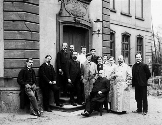 A black and white photograph of a group of people in front of a building, one person sits on a chair while others stand around him.