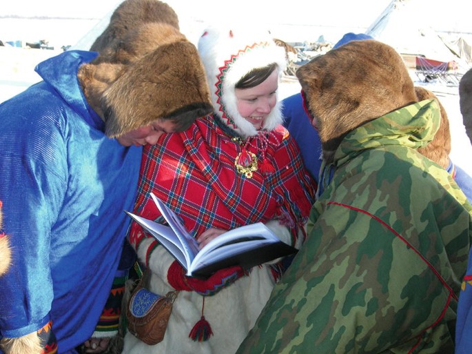 A photograph of 3 people wearing winter clothes and having conversation. The person in the middle holds a book in hand, while another person looks into that book.