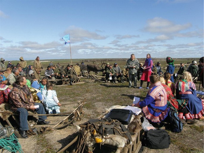 A photograph of a group of people sitting in a circular manner. Some people are in ethnic clothes. There are a couple of animals and a large grassland in the background. There are scattered clouds in the sky.