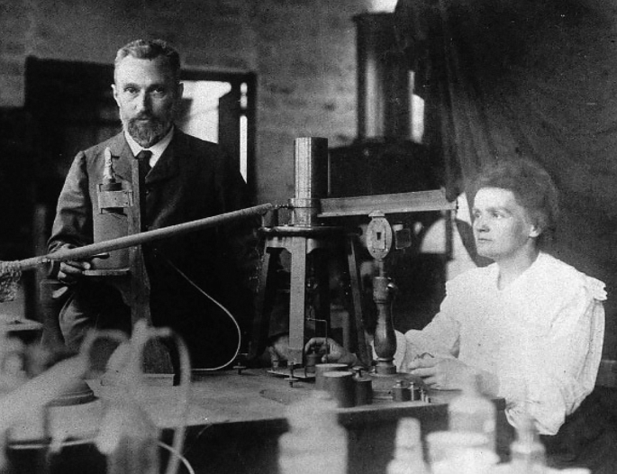 A photograph of Marie Curie and Pierre Curie in a laboratory. Marie Curie observes through a microscope.