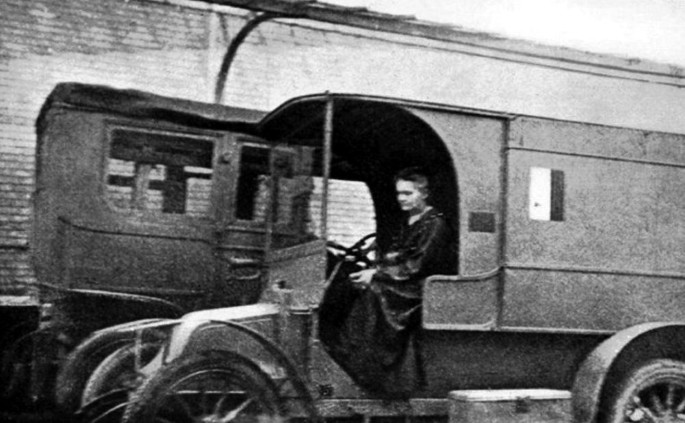 A photograph of Marie Curie sitting in the passenger seat of a motor vehicle. The back of the vehicle is closed.