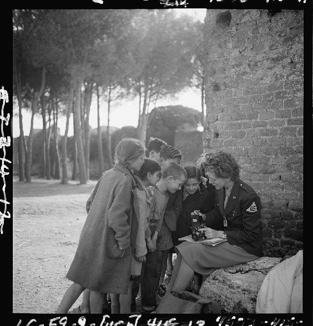 A photograph of a lady sitting in front of a wall holding a camera on her lap explains and shows something to the children standing around her. There are trees in the background.