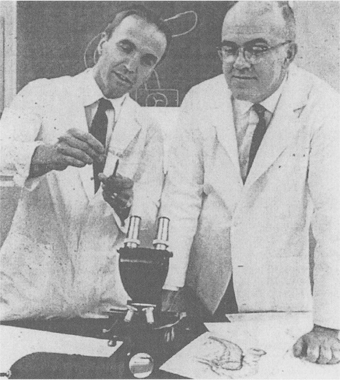 A grayscale picture of Richard Lillehei and William Kelly in a laboratory, looking at an object held and pointed by Richard Lillehei. A microscope and a few pieces of paper are on the table in front of them.