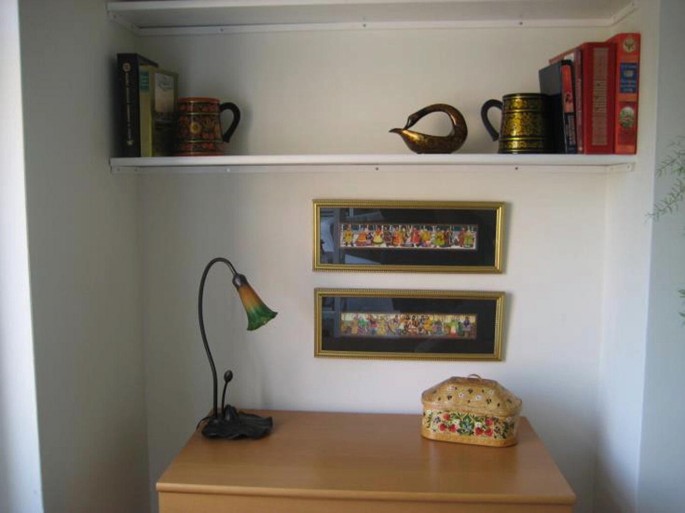 A photo of the home interior which contains a desk with a night lamp and a box is placed on the desk. Two photo frames hang on the wall and a shelf contains a few books and a few antique things.