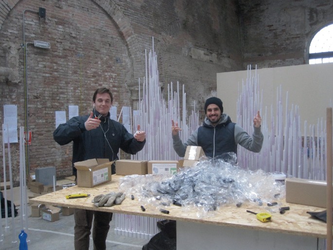 A photograph of 2 men who stand and pose in front of the till rolls sculpture. Package boxes, plastic covers, and tools are kept on a table in front of them.