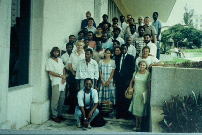 A photograph of a group of people who stands on wide stairs and looks straight into the camera. A person sits on the last stair. Trees, shrubs, and other plants are in the background.