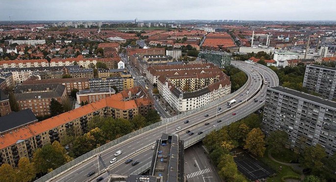 A photograph of a crowded city has a long overpass that bisects the city. There are large buildings on either side, both modern and traditional. There are many cars on the overpass. Spaces between the buildings are filled with trees, parks and tennis courts.