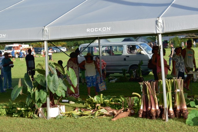A photograph of a tent with people, vegetables, and plants in it. A few vehicles are in the background.