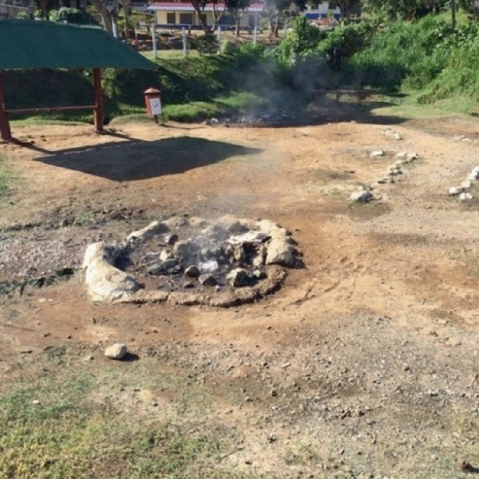A photograph of a hot spring in the ground. The area is damp and is surrounded by small rocks. Smoke is coming out of it.