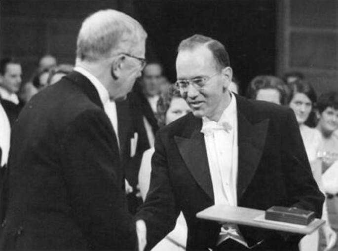 A photo. In the foreground, 2 men in suits, look at each other and shake hands. The man on the right, faces the camera and holds a case placed on a tray. The one on the left turns away from the camera and towards the other man. Many rows of seated audience members appear in the background.