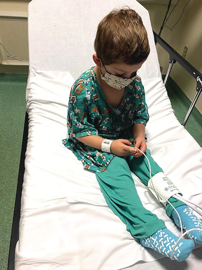 A photograph of a sick boy with a mask sitting on a hospital bed. 2 tubes are connected from his right-hand finger and from his left leg.