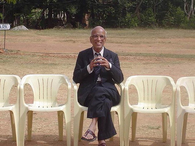 A photograph of Kanakanahalli Ramachandra. He is dressed formally in a suit and slippers and sits in the middle chair, with two empty chairs on either side, and trees in the background.