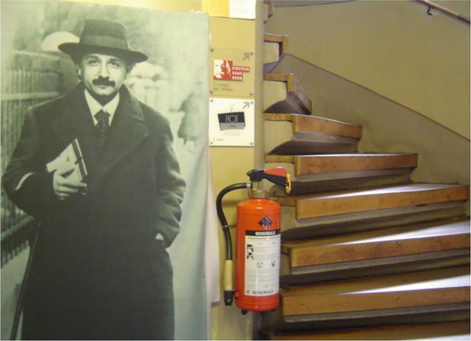 A photo of Einstein's home. A photo of Einstein is placed on the wall to the left. A curved staircase is to the right. A fire extinguisher is between the photo and staircase.