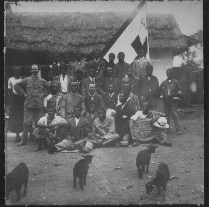 A monochrome photograph of a group of people in front of a village house with a thatched roof. Some of them sit, while others stand behind. A flag with a cross symbol is behind them. 4 stray dogs are on the road in front of them.