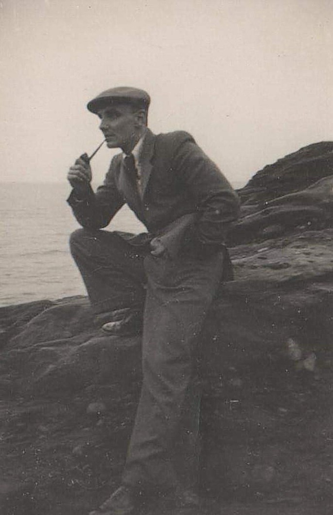 A photograph of David sitting on a rock with a smoking pipe placed in his mouth.