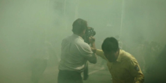 A photograph of a man holding up a large video camera. The environment is covered in a shroud of mist. Another man holds onto the forearm of the man with the camera while looking away,