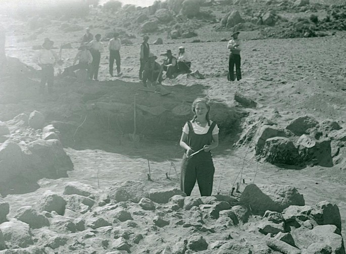 A greyscale image of a field with a woman in the foreground and a few other people in the background.