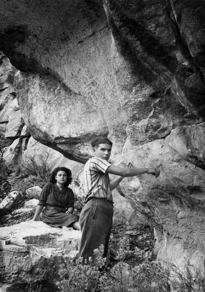 Two monochrome photos of a few people outside a rock structure. The top photo has a boy and a girl. The photo at the bottom has a man, a boy, and a girl.
