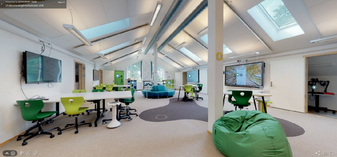 A photograph of a classroom furnished with tables and chairs on wheels, as well as a sofa, a couch, and smart televisions. A rack is in the corner of the room. The ceiling of the classroom is made of glass.