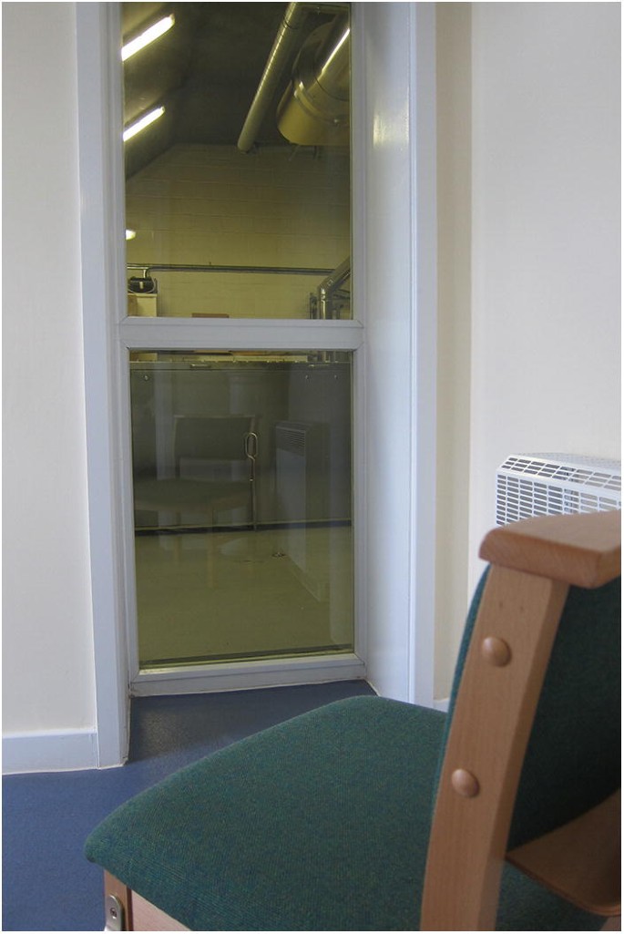 A photograph of the cremator viewing room with a chair. The chair is towards the viewing glass sheet of the cremation chamber.