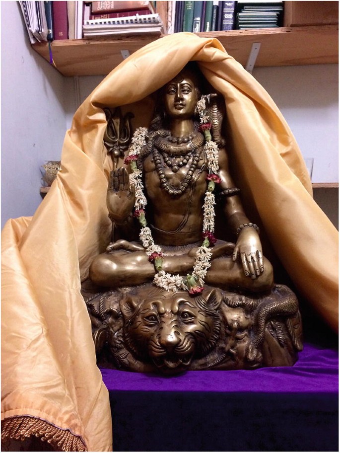 A photograph of a Shiva statue. There is a flower garland around the neck of the statue. The top of the statue is covered with fabric.